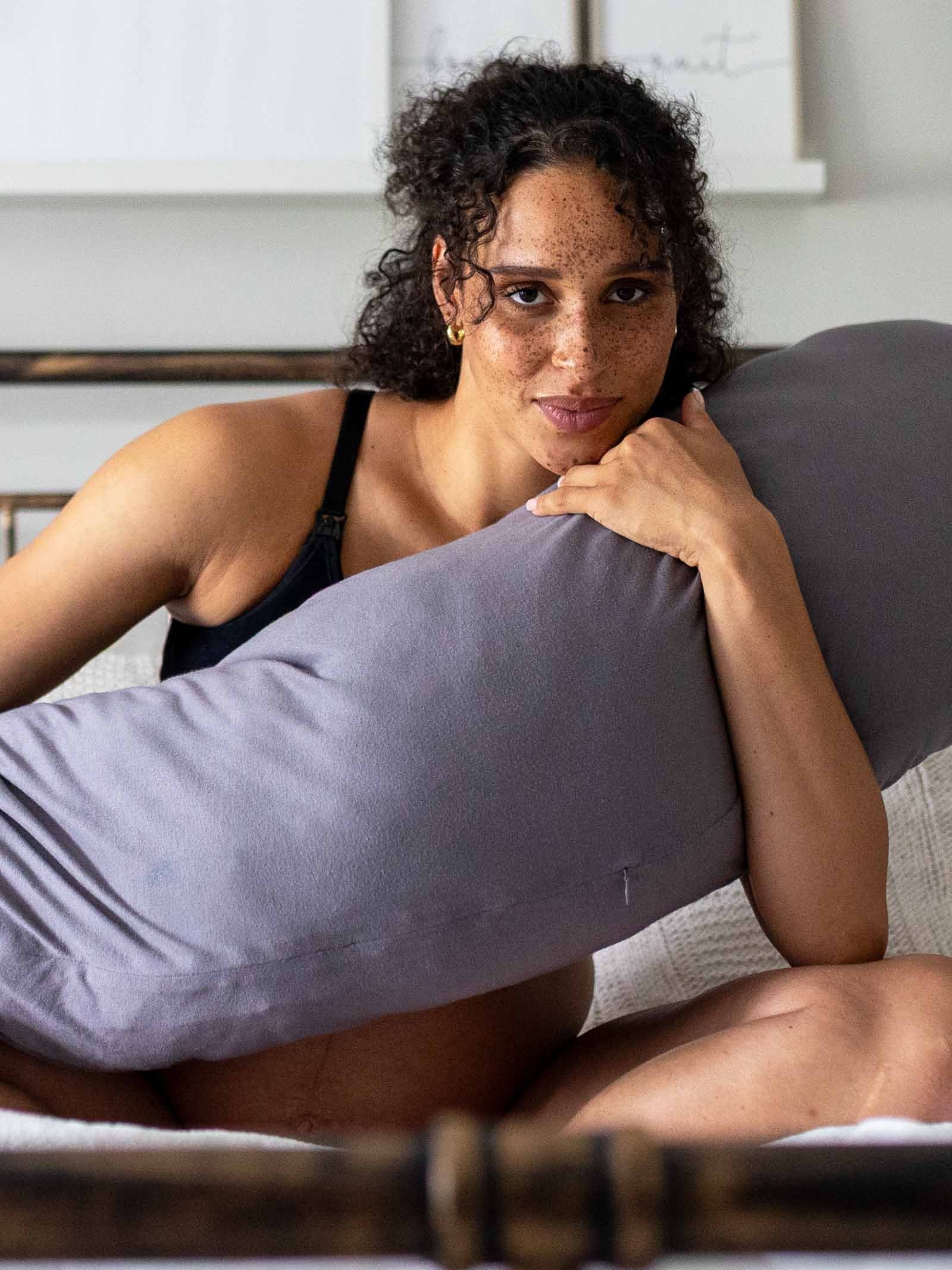 A woman sitting on a bed, hugging the grey pregnancy pillow.