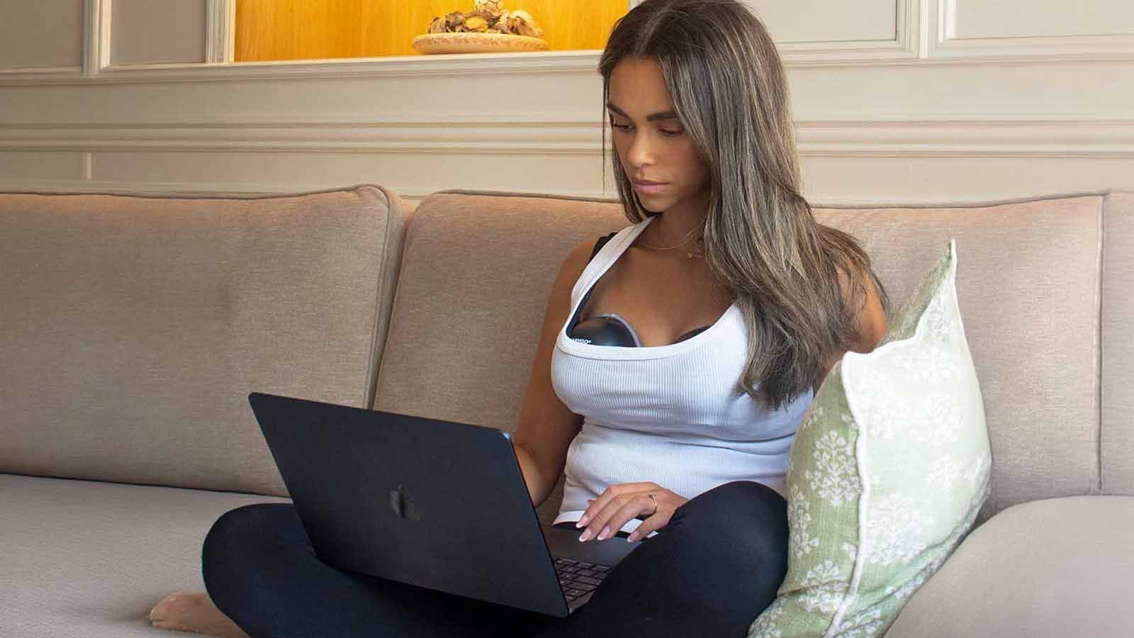 A mother working on a laptop on a sofa, the pump inside her top.